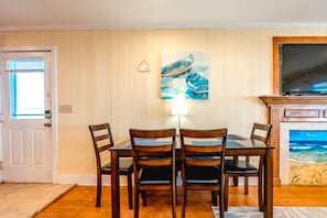 A dining area with a table and chairs, a painting above the table, and a fireplace with a painting on the wall. - Good Company - Classic 3BR Oceanfront Beach Cottage with Screen Porch (Topsail Beach)
