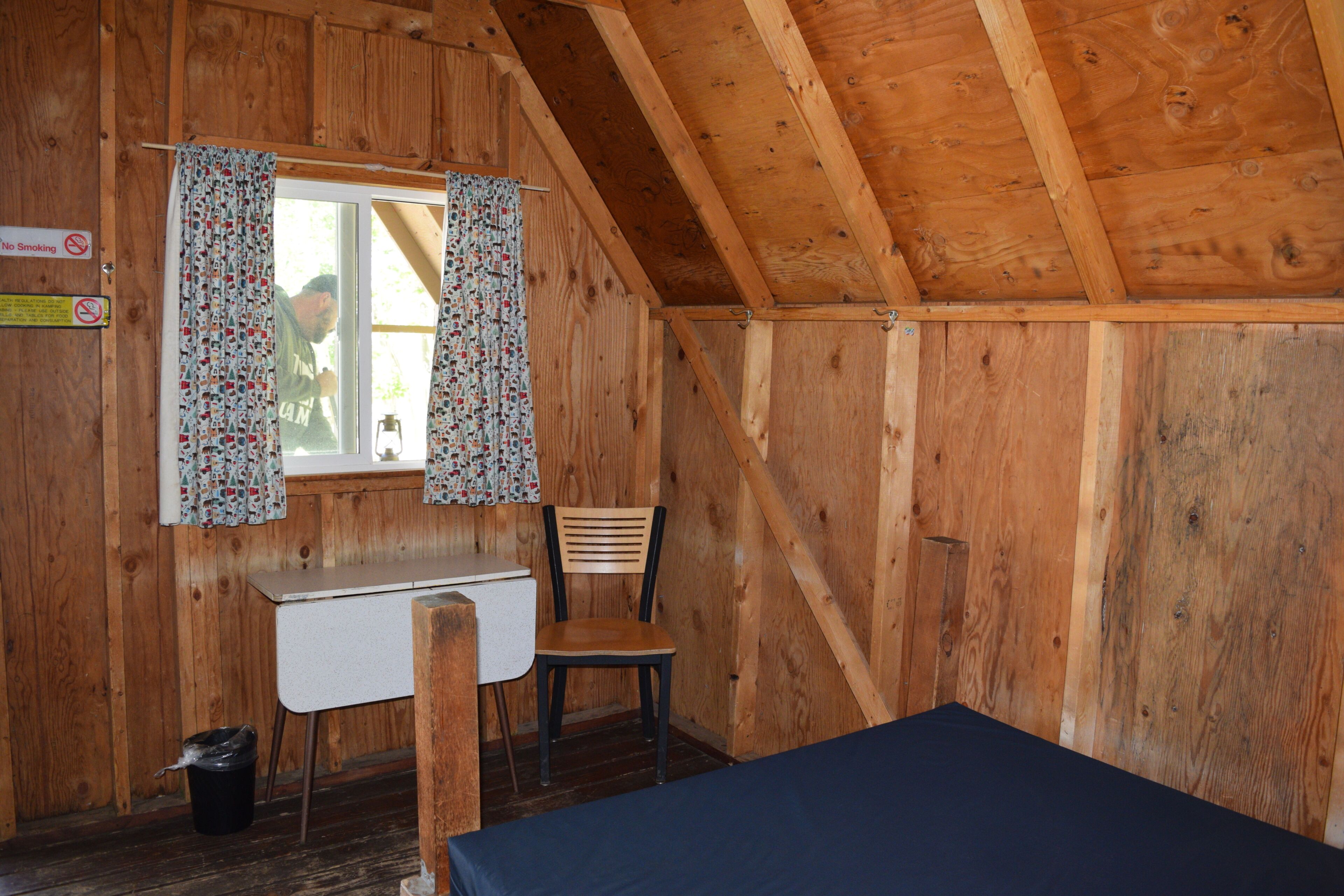 A small cabin with a wooden interior, a single window with curtains, a desk, a chair, and a bed.