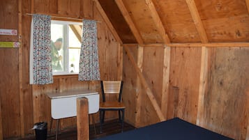 A small cabin with a wooden interior, a single window with curtains, a desk, a chair, and a bed.