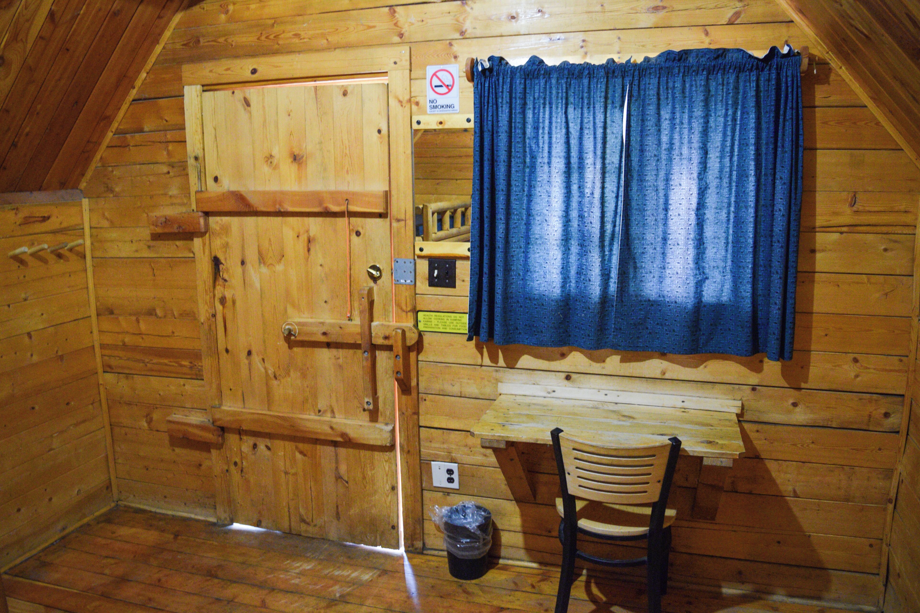 Una habitación de madera con una puerta, un banco, una silla y una ventana con cortinas.