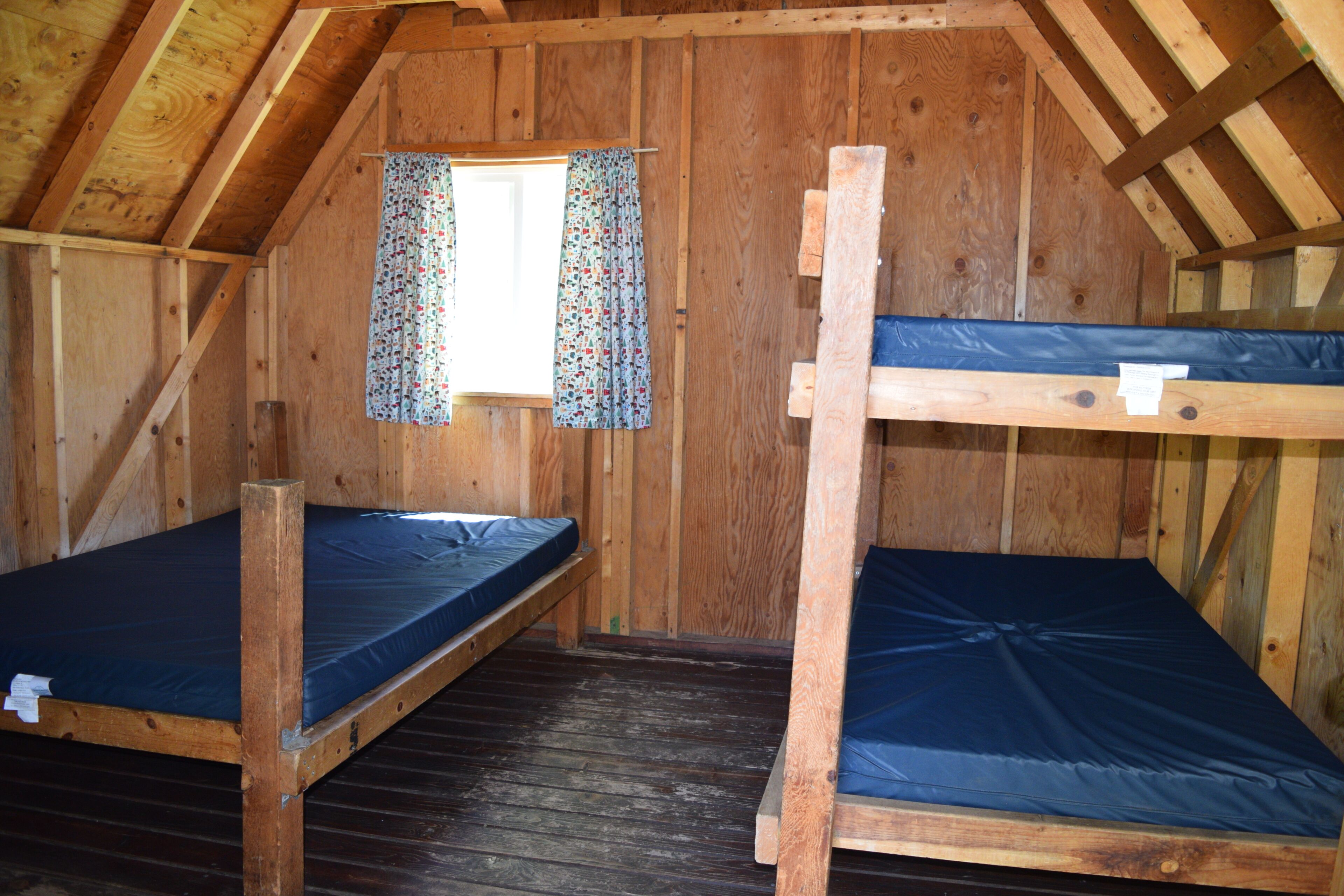 A wooden cabin with two bunk beds, a window with curtains, and a sloped ceiling.