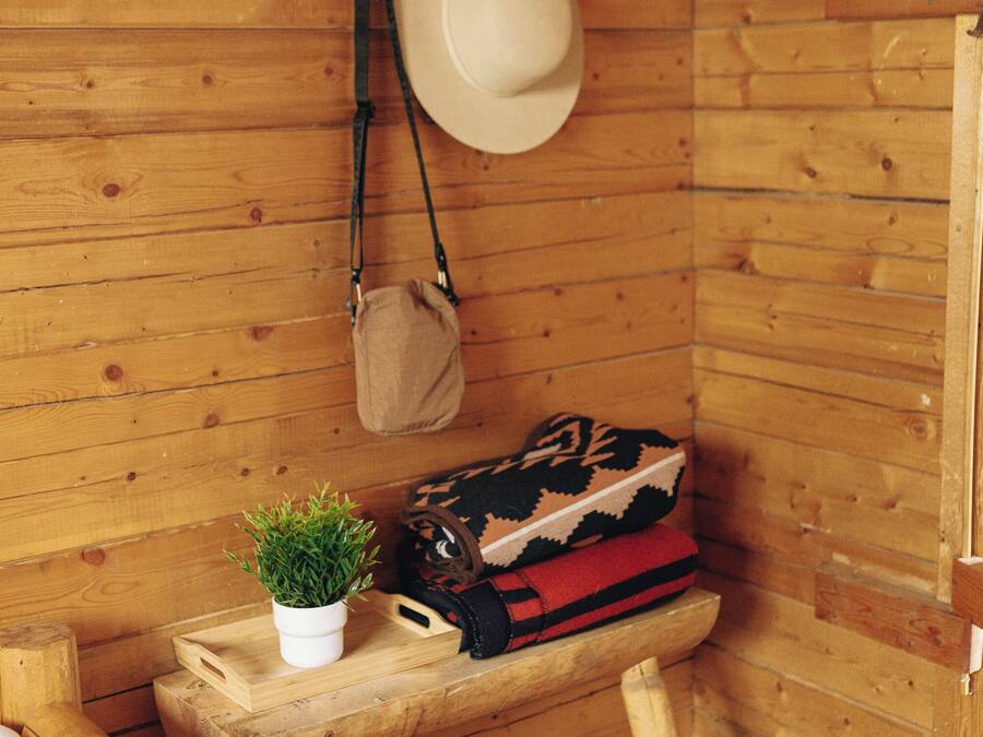 A wooden cabin interior with a bed, a small table with a plant, a hat, and a bag.