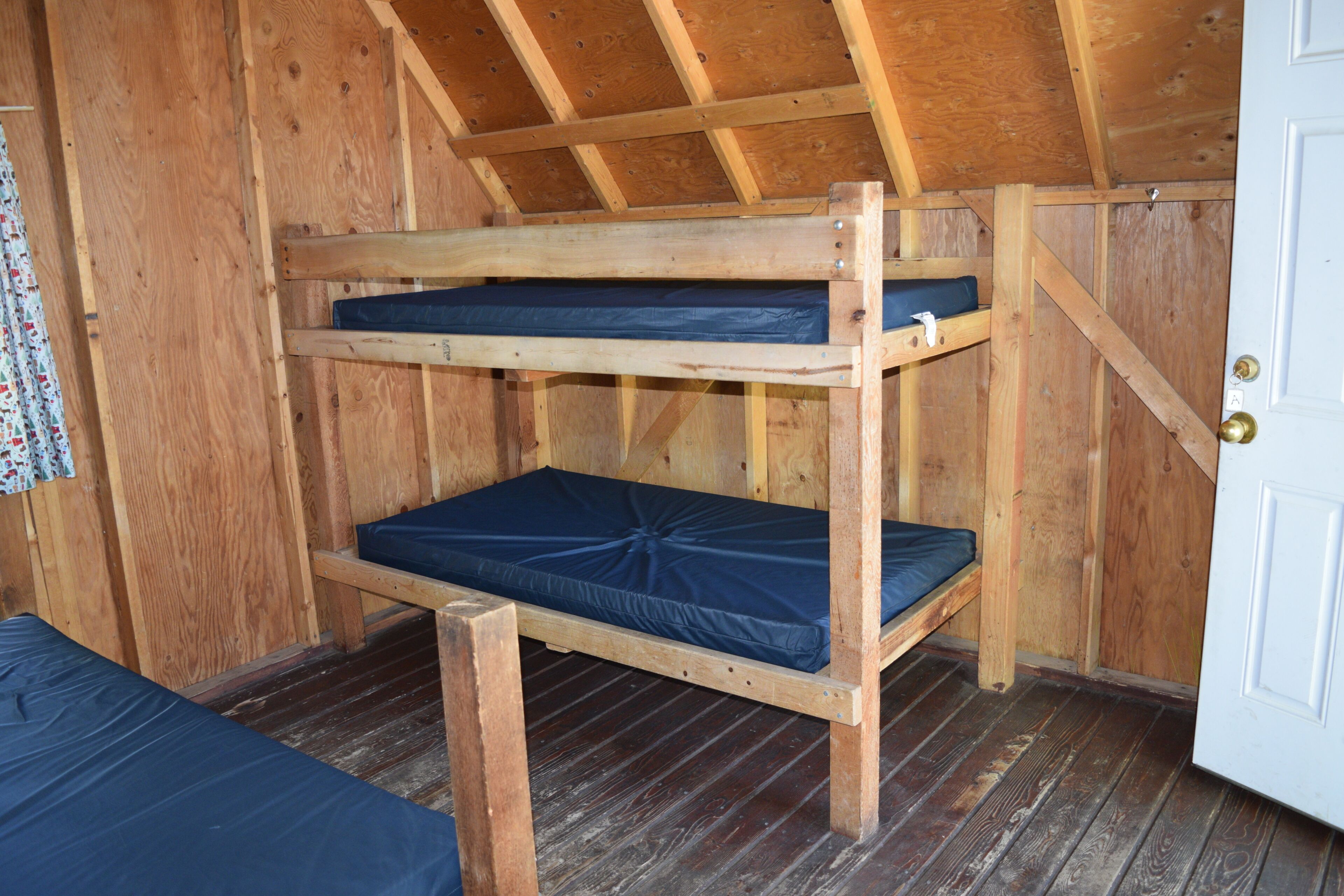 Een stapelbed met houten frame en blauwe matrasbeschermers in een kamer met houten wanden en een schuin dak.