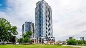 A modern high-rise building with a glass facade, surrounded by a green park and other buildings in the background. - Newly Built Modern Condo With Stunning City Views (Kitchener)