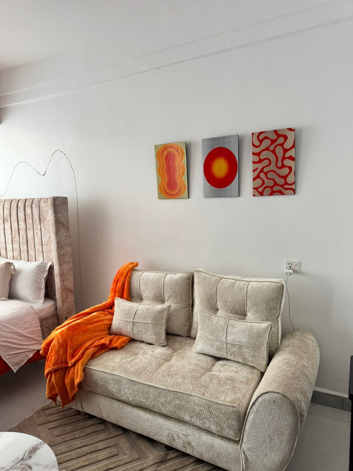 Een moderne woonkamer met een beige bank, een kamerkleed met dessin en drie abstracte schilderijen aan de muur.