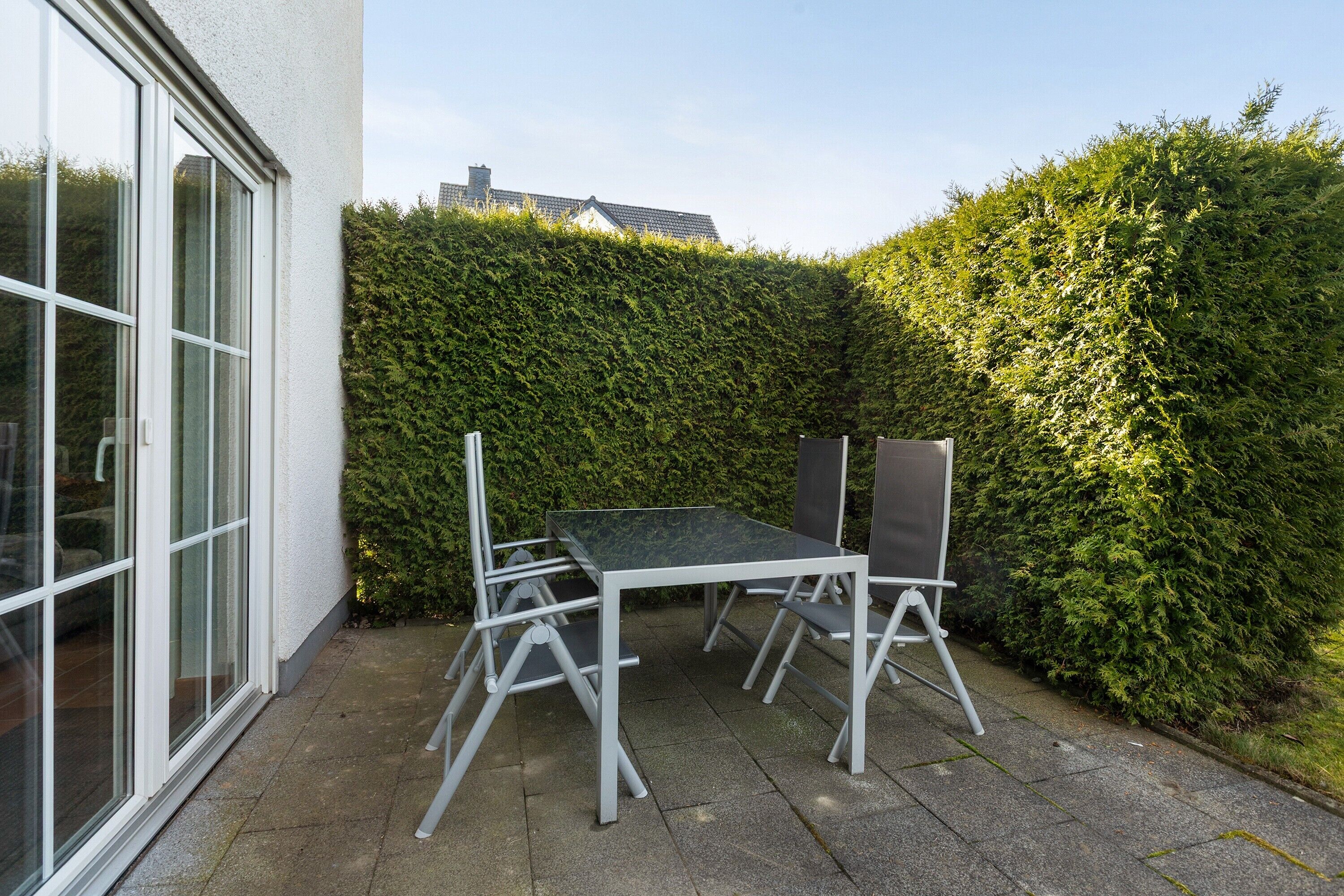A patio with a table and chairs, a hedge, and a building with large windows.