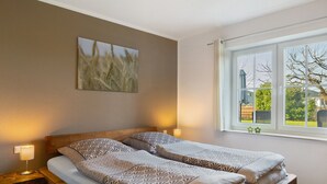 Una cama doble con ropa de cama estampada, una mesita de noche con lámpara, una ventana con vistas y un cuadro de un campo de trigo en la pared.