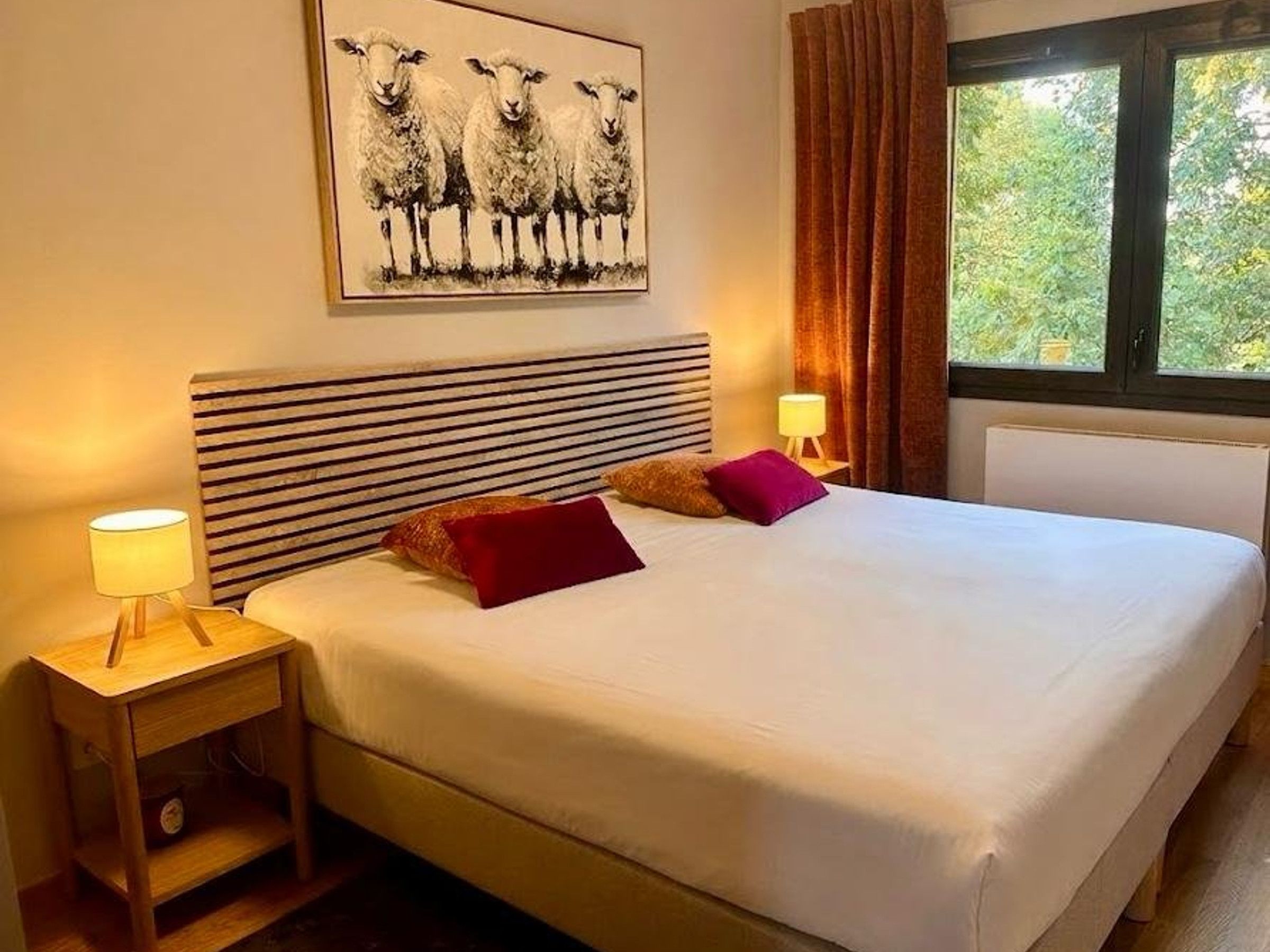 A hotel room with a large bed, two bedside lamps, a headboard with horizontal stripes, a window with curtains, and a painting of sheep on the wall.