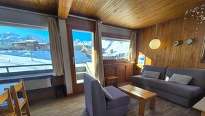 Ein gemütliches Zimmer mit Holzbalkendecke, einem grauen Sofa, einem kleinen Holztisch und Blick auf einen verschneiten Berg.