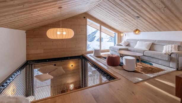 En modern inredning med ett sluttande tak, en sittgrupp med soffa och kuddar, ett litet bord och ett stort fönster som erbjuder utsikt över omgivningen.