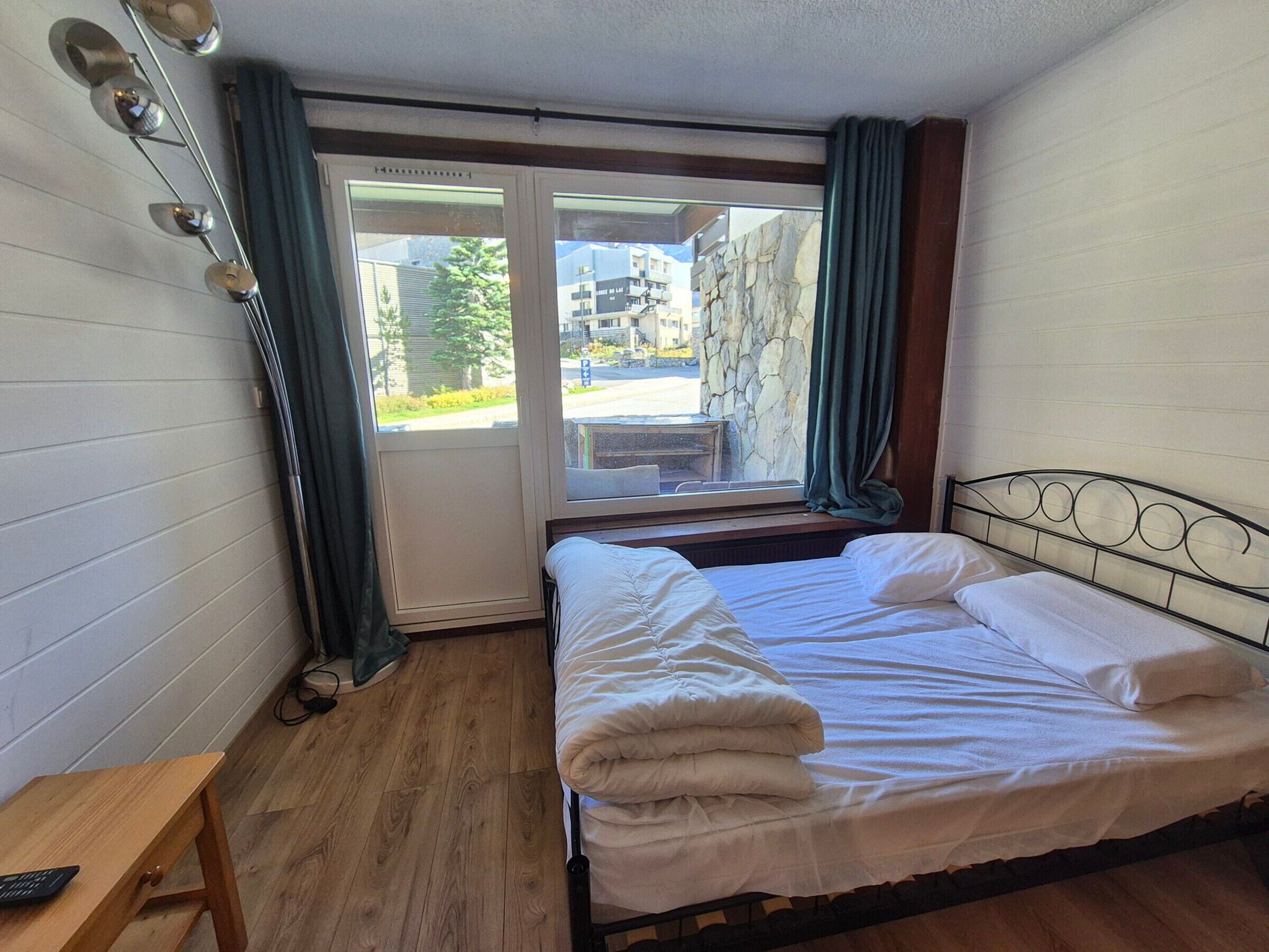 Quarto com cama, piso de madeira, janela com cortinas e vista para prédios.