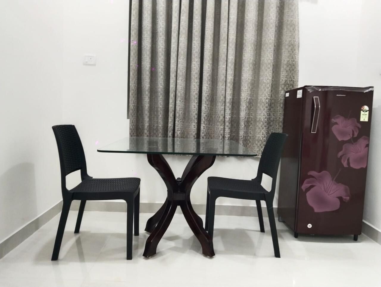 A dining area with a glass-top table and two chairs, a refrigerator with floral design, and a window with curtains.