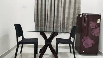 A dining area with a glass-top table and two chairs, a refrigerator with floral design, and a window with curtains.