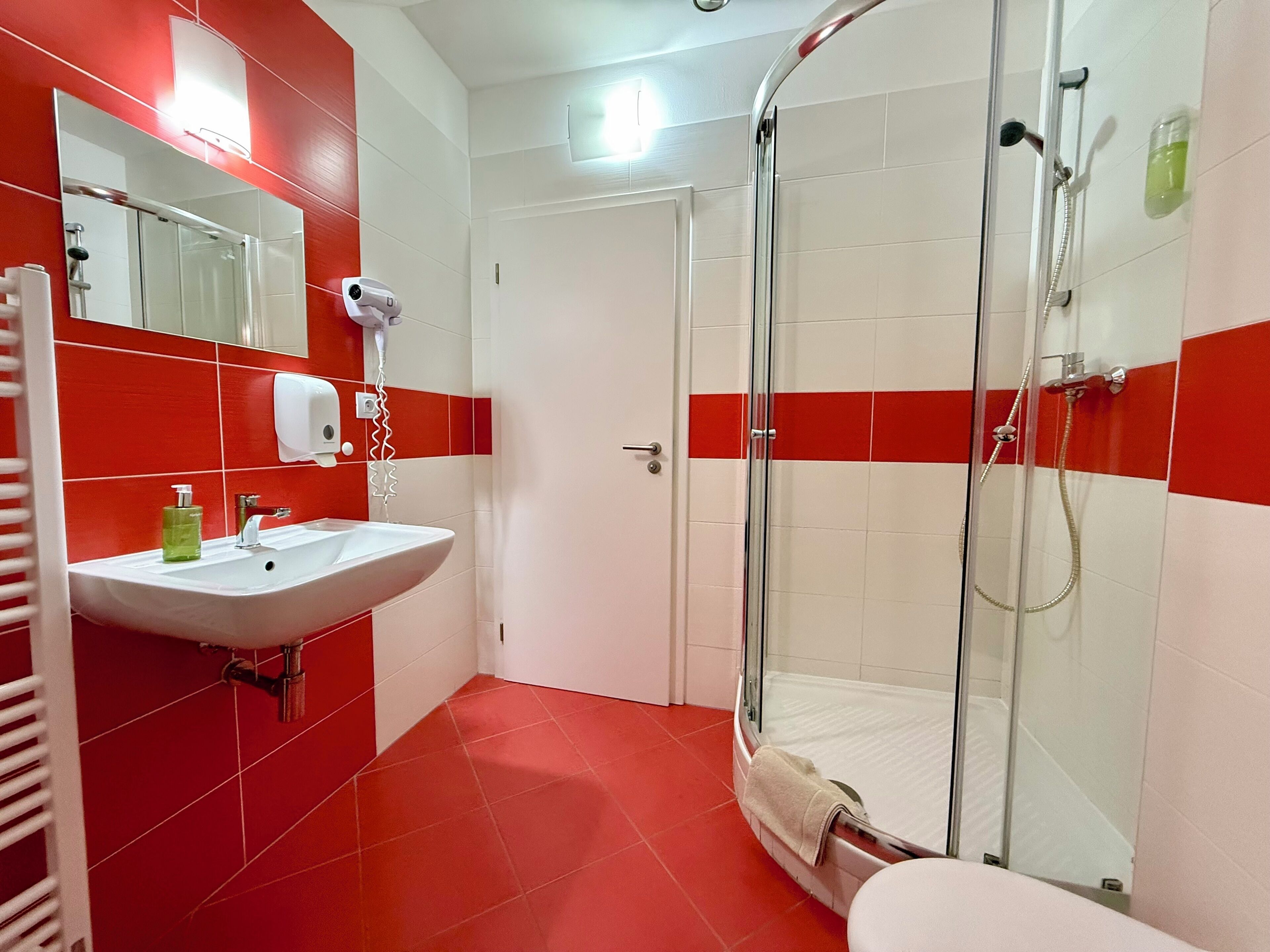 Un baño con ducha envuelta en vidrio, un lavamanos blanco y paredes con azulejos rojos.