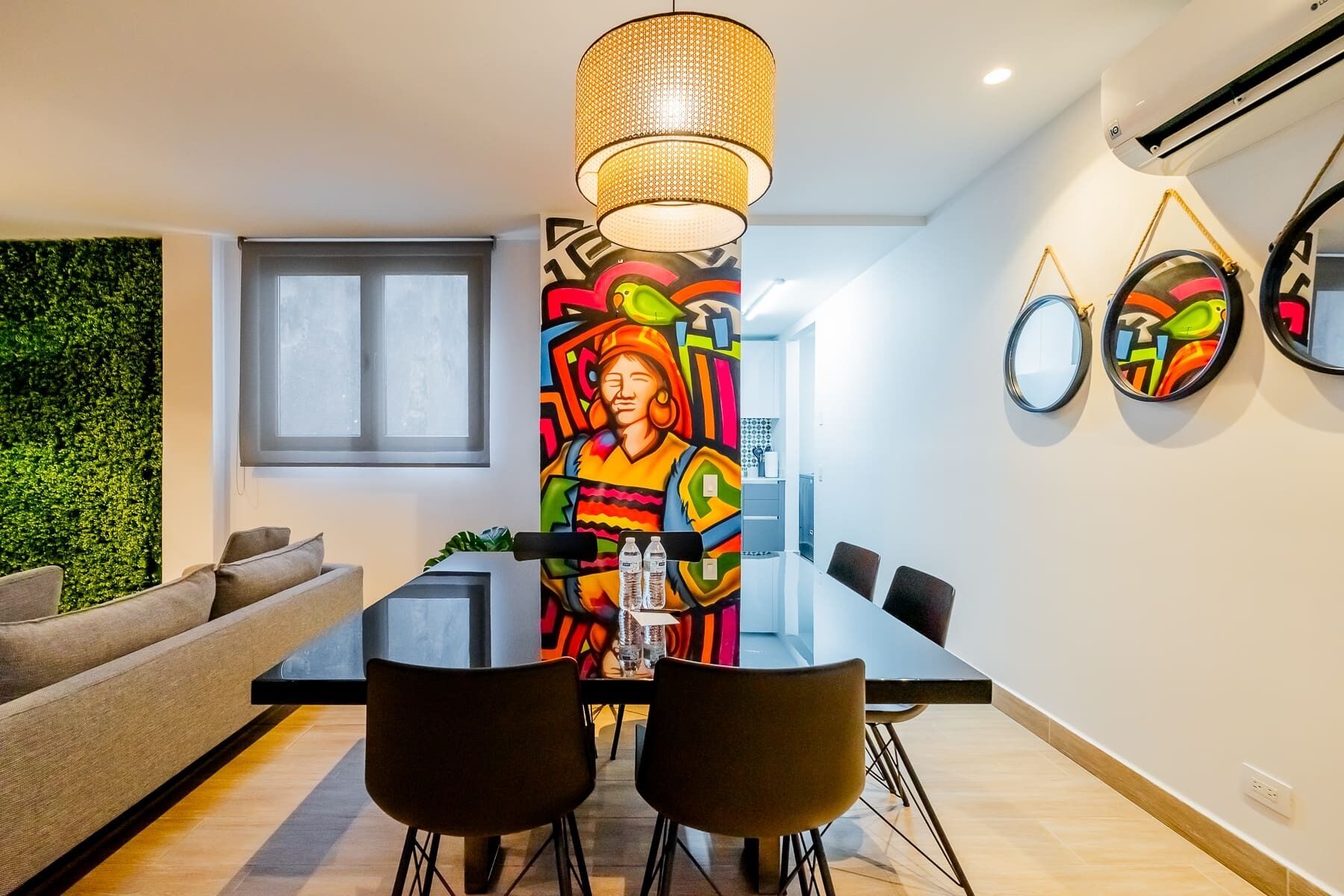 Un espace de salle à manger moderne avec une table en verre, des chaises noires, un tableau abstrait aux couleurs vives, un luminaire suspendu et un mur vert.