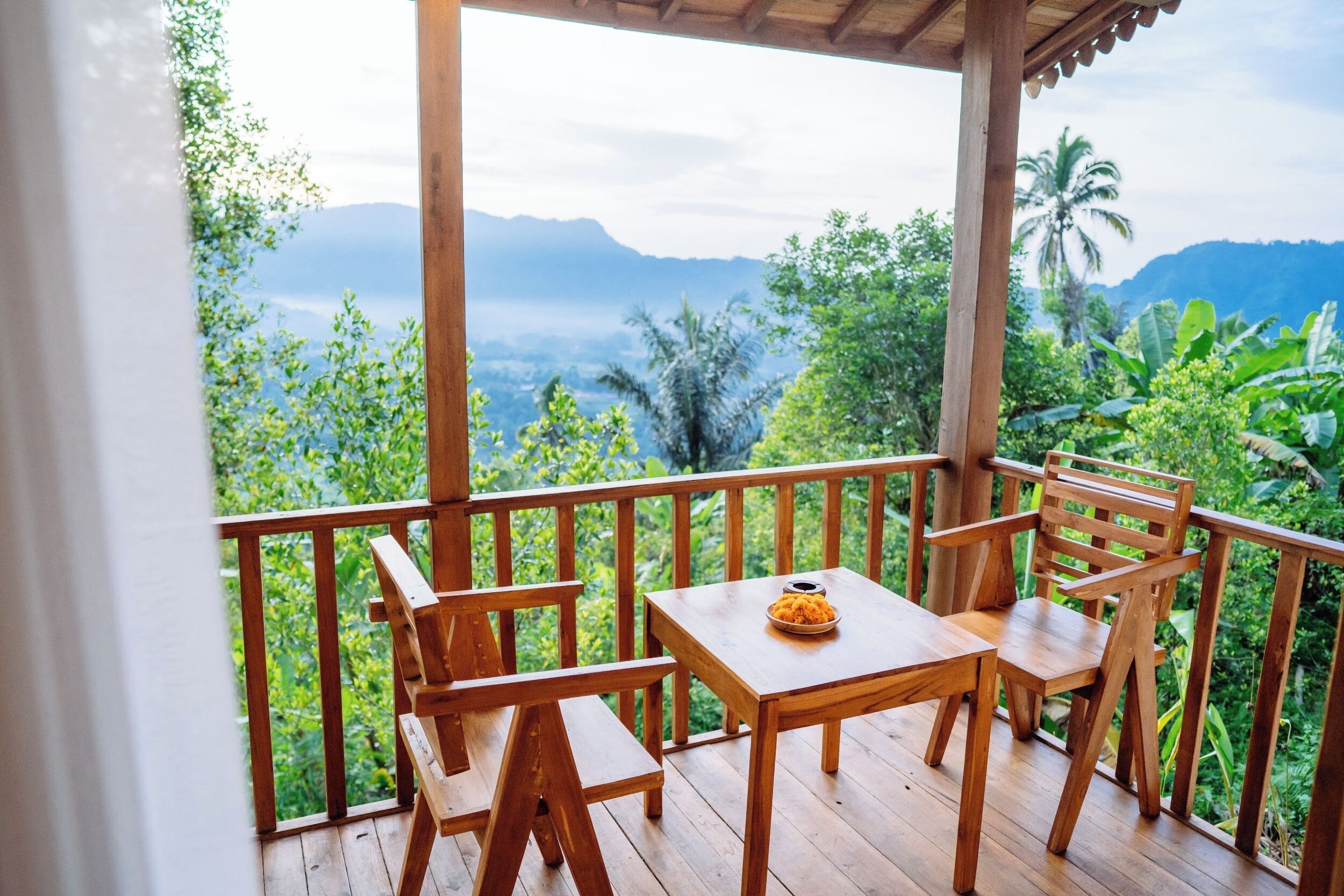 Una terraza de madera con una mesa y sillas que da a un paisaje exuberante con montañas y vegetación tropical.