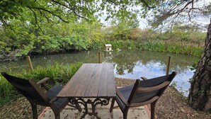 Una mesa de madera con dos sillas en un jardín que tiene un estanque y un comedero para pájaros al fondo.