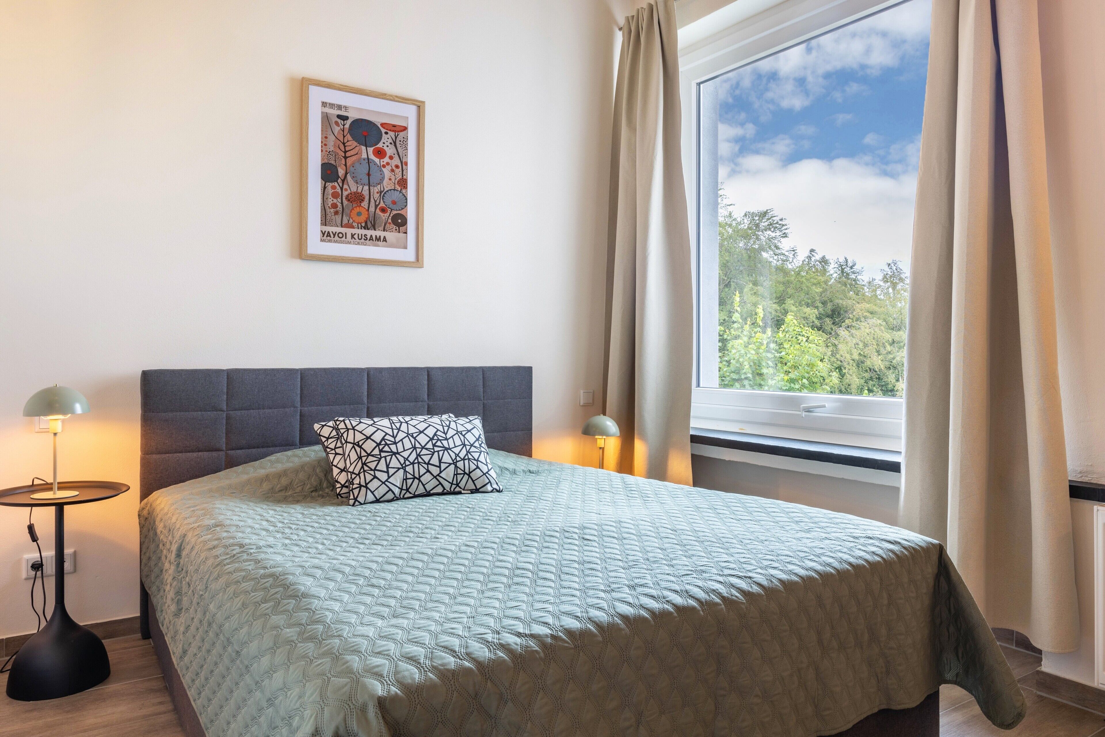 Una cama bien tendida con una colcha estampada, una obra de arte enmarcada encima y una ventana con cortinas.