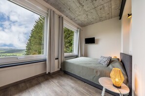 Une chambre avec une grande fenêtre, un lit recouvert d’une couvre-lit à motifs, une lampe de chevet et un téléviseur à écran plat fixé au mur.