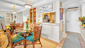 A dining area with a round glass table, wicker chairs, and a kitchen with wooden cabinets and a microwave. - Oceanfront Royal Mauian #515 - 2BR/2BA with Panoramic Views Near Kamaole Beaches (Kihei, Maui)