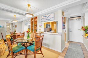 A dining area with a round glass table, wicker chairs, and a kitchen with wooden cabinets and a microwave. - Oceanfront Royal Mauian #515 - 2BR/2BA with Panoramic Views Near Kamaole Beaches (Kihei, Maui)