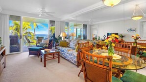 A spacious living room with a large glass table, wicker furniture, and a view of palm trees through the glass doors. - Oceanfront Royal Mauian #515 - 2BR/2BA with Panoramic Views Near Kamaole Beaches (Kihei, Maui)
