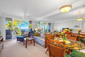 A spacious living room with a large glass table, wicker furniture, and a view of palm trees through the glass doors. - Oceanfront Royal Mauian #515 - 2BR/2BA with Panoramic Views Near Kamaole Beaches (Kihei, Maui)