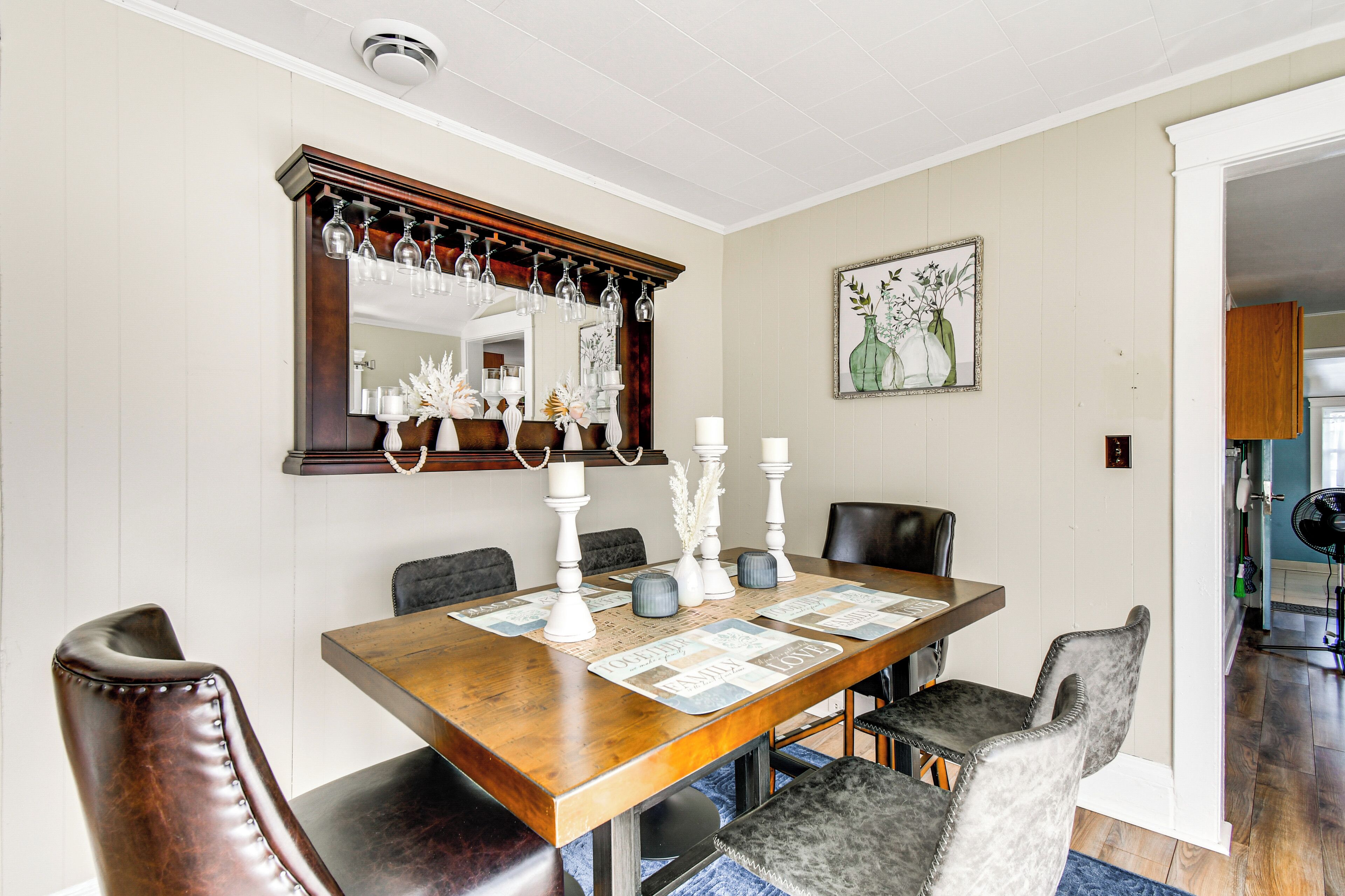 Un comedor con una mesa de madera, sillas tapizadas en negro y gris, un espejo grande con luces colgantes y un cuadro enmarcado en la pared.