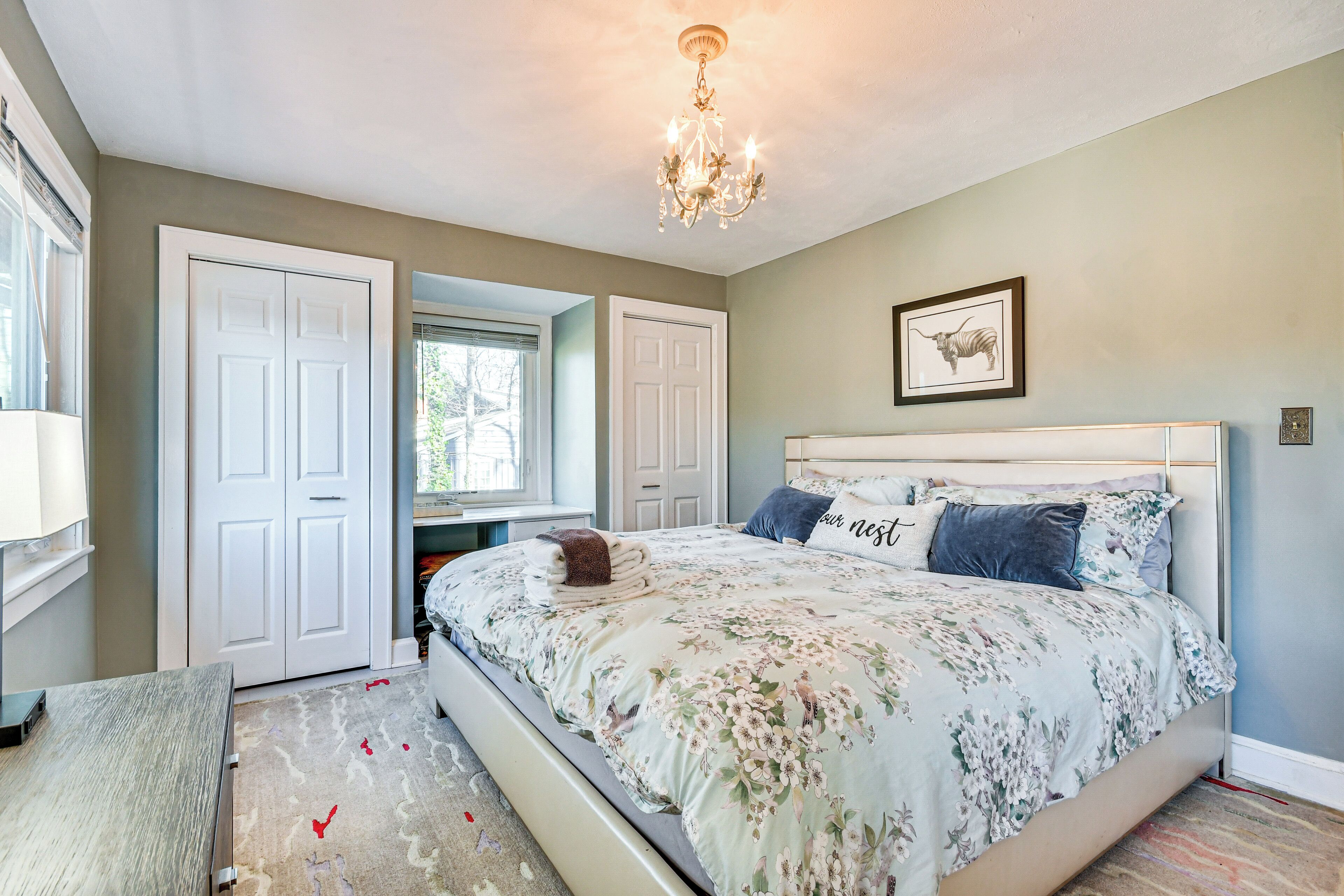 A bedroom with a large bed, two white doors, a chandelier, and a framed picture on the wall.