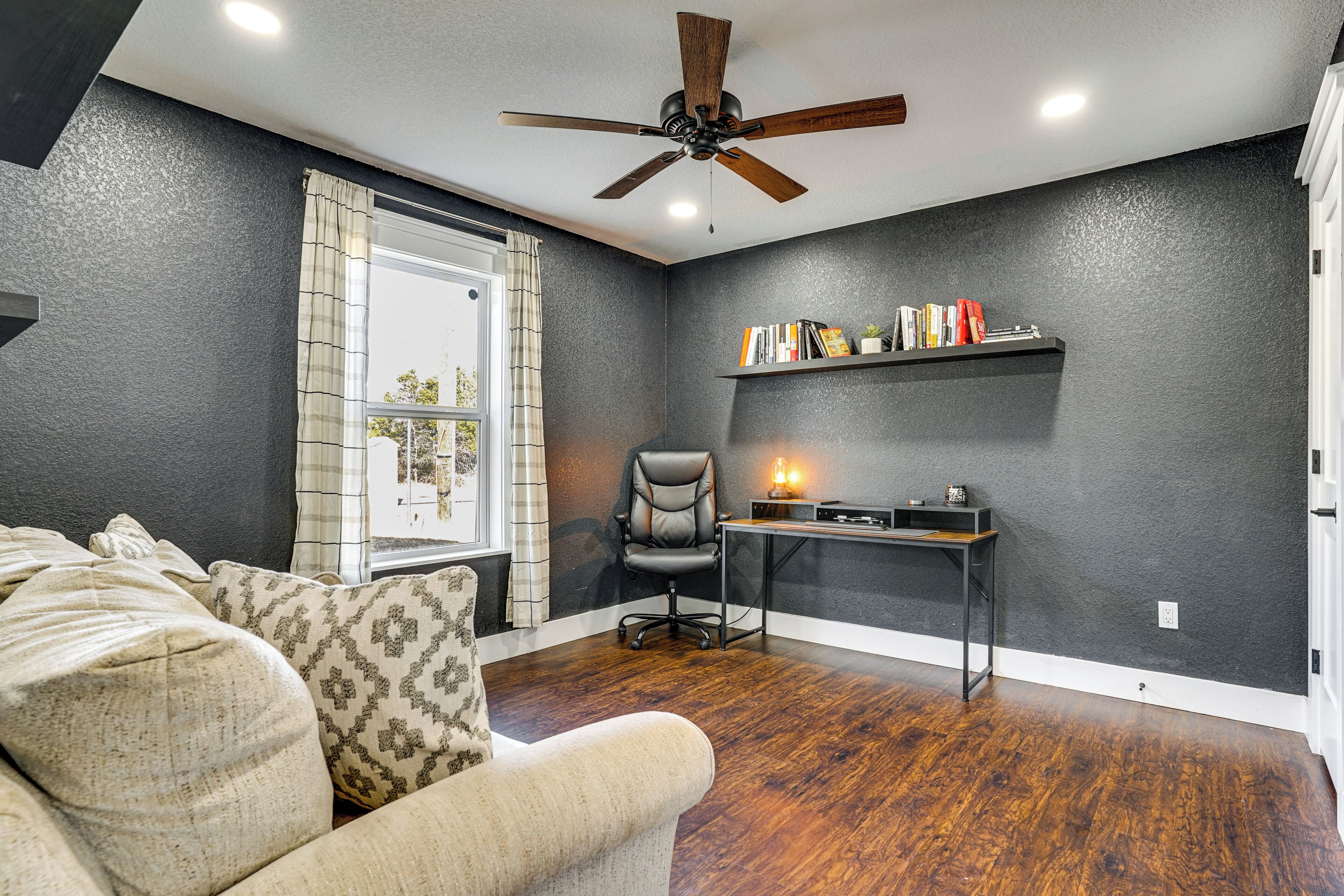 A room with a ceiling fan, a desk with a lamp, a chair, and a couch with patterned cushions.