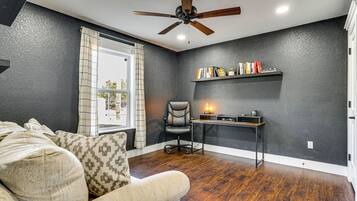 A room with a ceiling fan, a desk with a lamp, a chair, and a couch with patterned cushions.