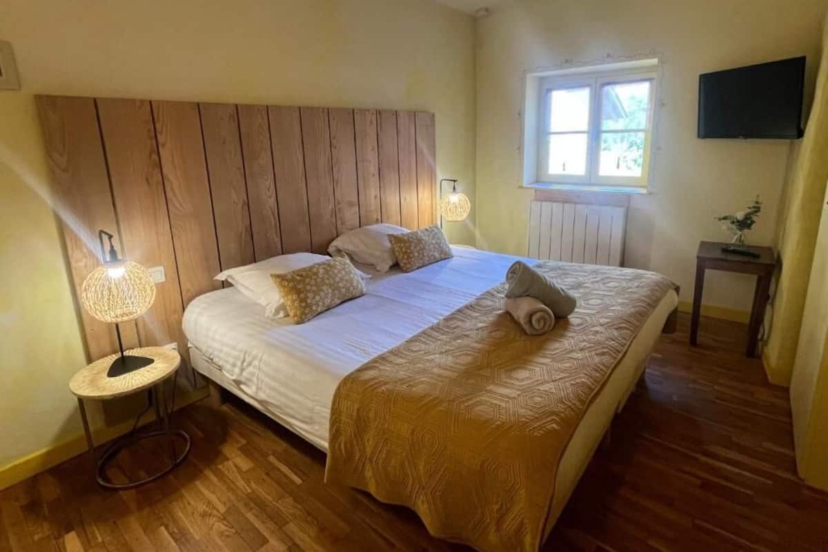Un dormitorio con cabecera de madera, cama con colcha estampada, lámpara de noche y ventana con vistas.