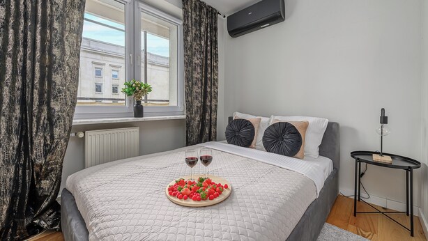 A bedroom with a bed, a bedside table, a window with curtains, and a radiator. - Lumina lux apart in the center of Warsaw (Warsaw)
