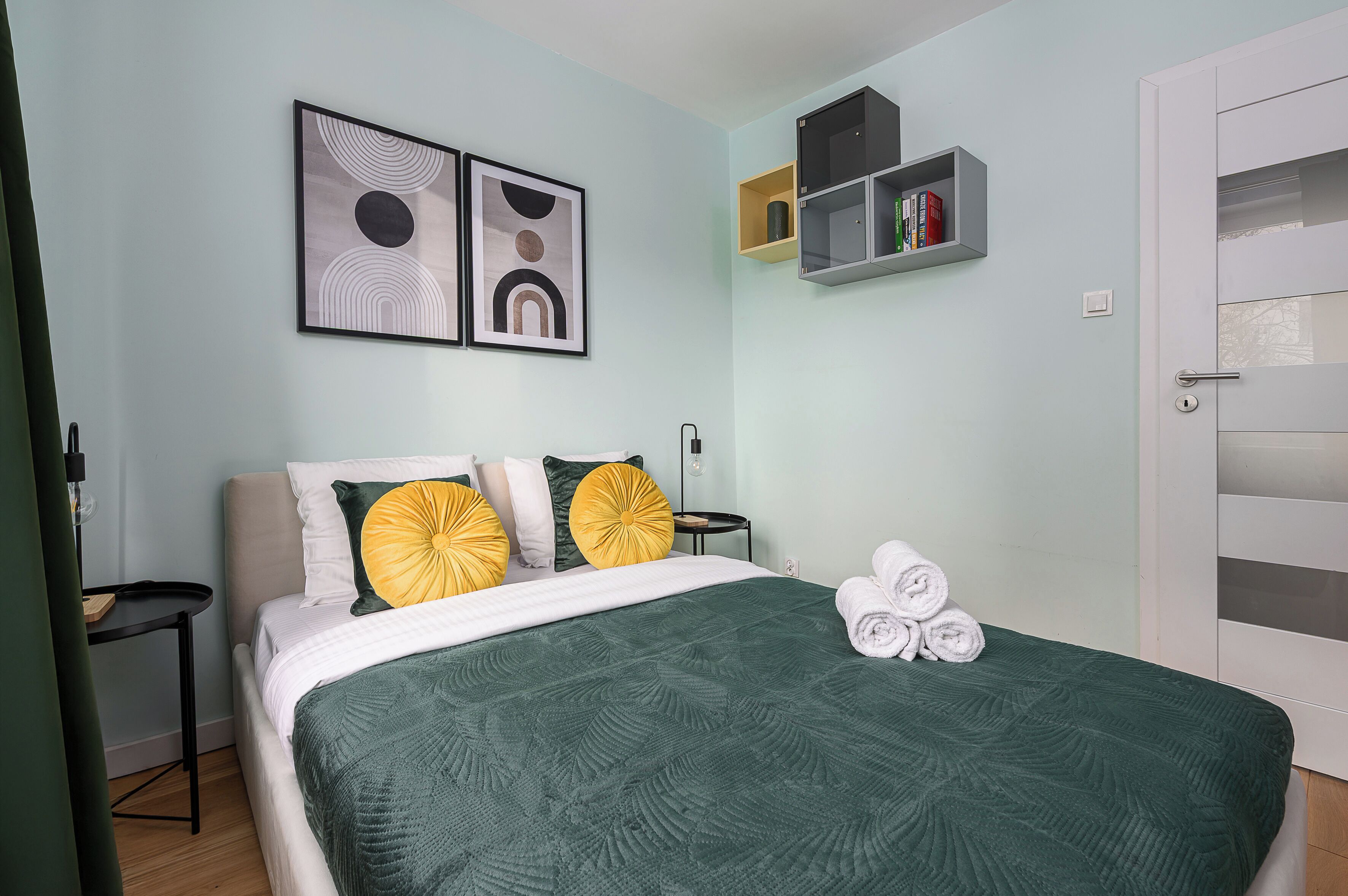 Een net opgemaakt bed met een groene sprei en gele kussens, een klein nachtkastje en een wandplank met boeken en een lamp.