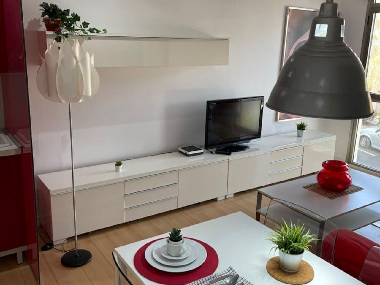 A modern living room with a flat-screen TV on a white cabinet, a floor lamp, and a dining area with a red table and white chairs.