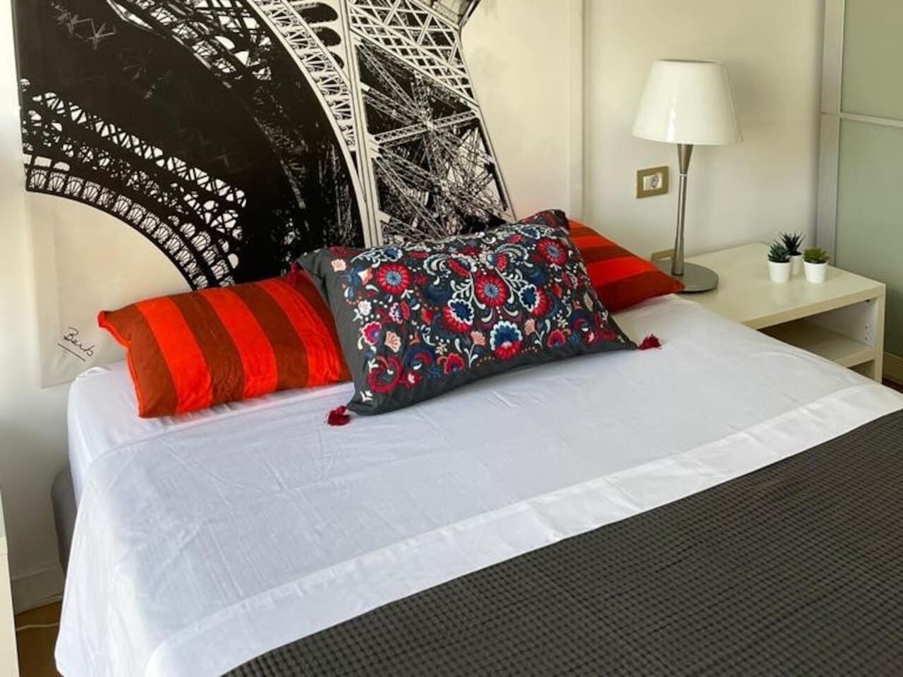 A neatly made bed with a white bedspread and decorative pillows, a bedside lamp, and a wall with a large black and white artwork.