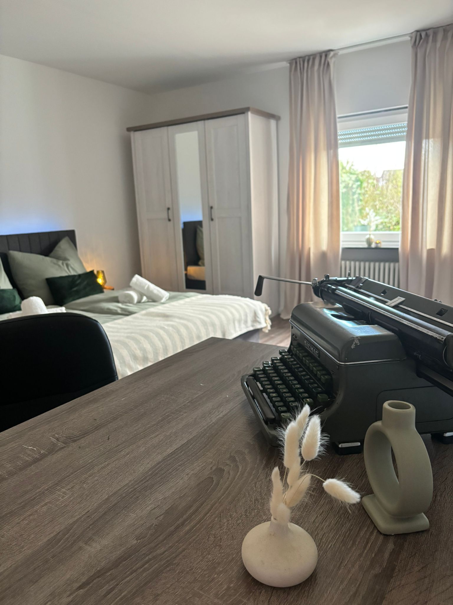 Ein Hotelzimmer mit einem Bett, einem Schreibtisch mit Schreibmaschine, einer Lampe und Blick auf Grün.