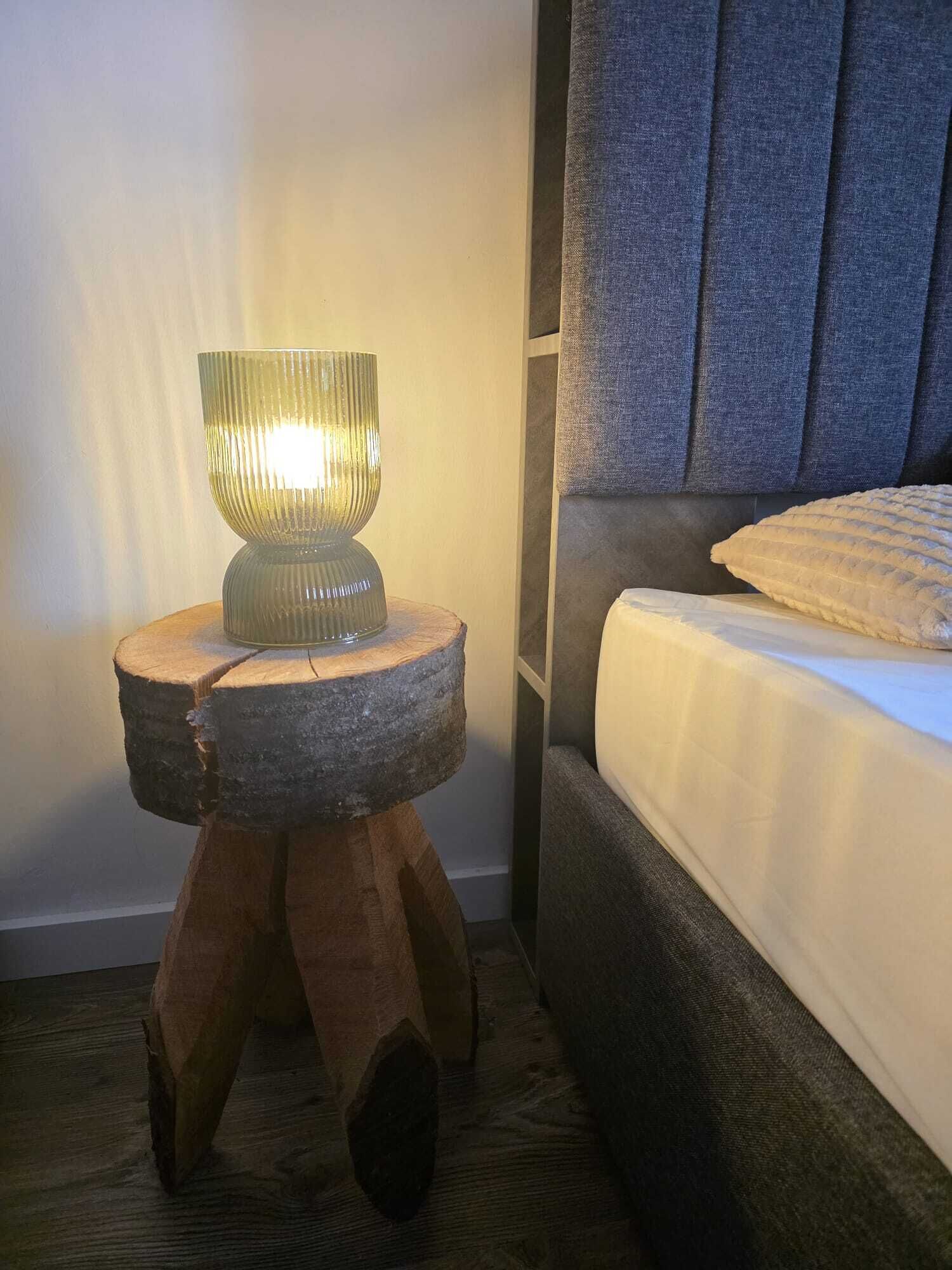 A modern bedroom with a wooden nightstand holding a glass lamp, a bed with a dark headboard, and a textured throw pillow.