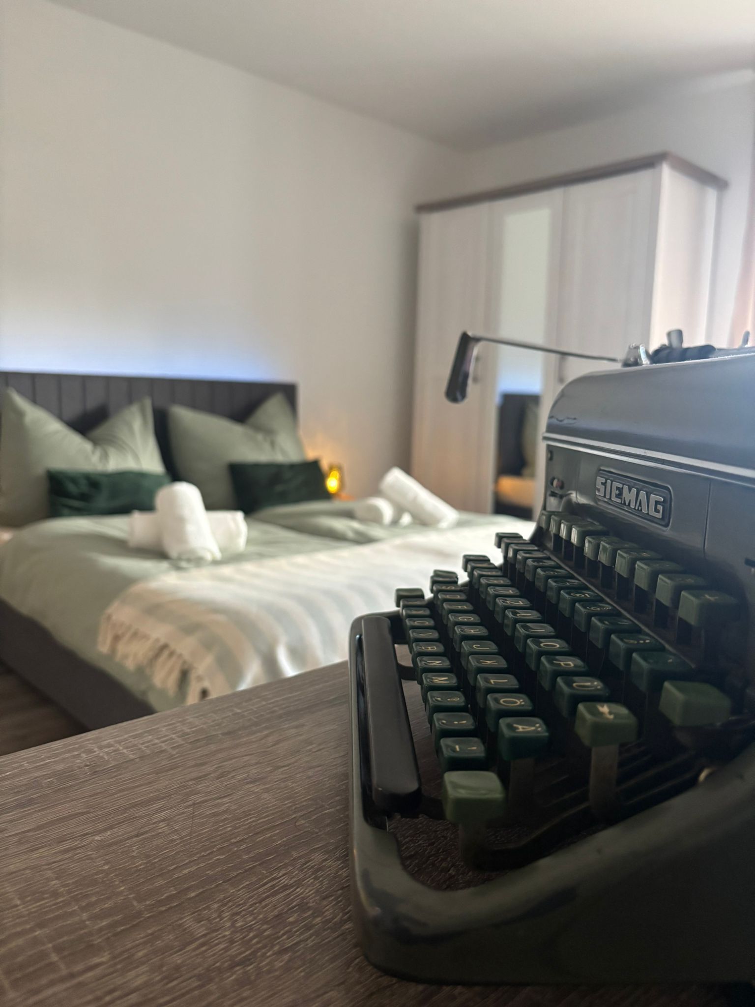 Una máquina de escribir antigua sobre una mesa de madera en una habitación de hotel con cama, almohadas y mesita de noche.