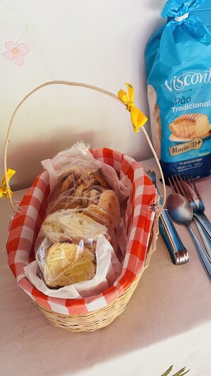 A basket of bread wrapped in plastic, a blue bag of bread, and a fork and spoon on a table. - Vale das Rainhas Pousada Santo Amaro BA (Santo Amaro)