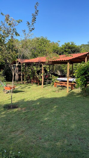 Un espacio al aire libre rústico con una pérgola de madera, una mesa con mantel a cuadros y una carretilla.