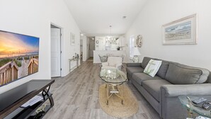 Ein modernes Wohnzimmer mit einem grauen Sofa, einem Glastisch und einem Fernseher auf einem schwarzen Standfuß. Ein Holzboden und ein Deckenventilator sind zu sehen.
