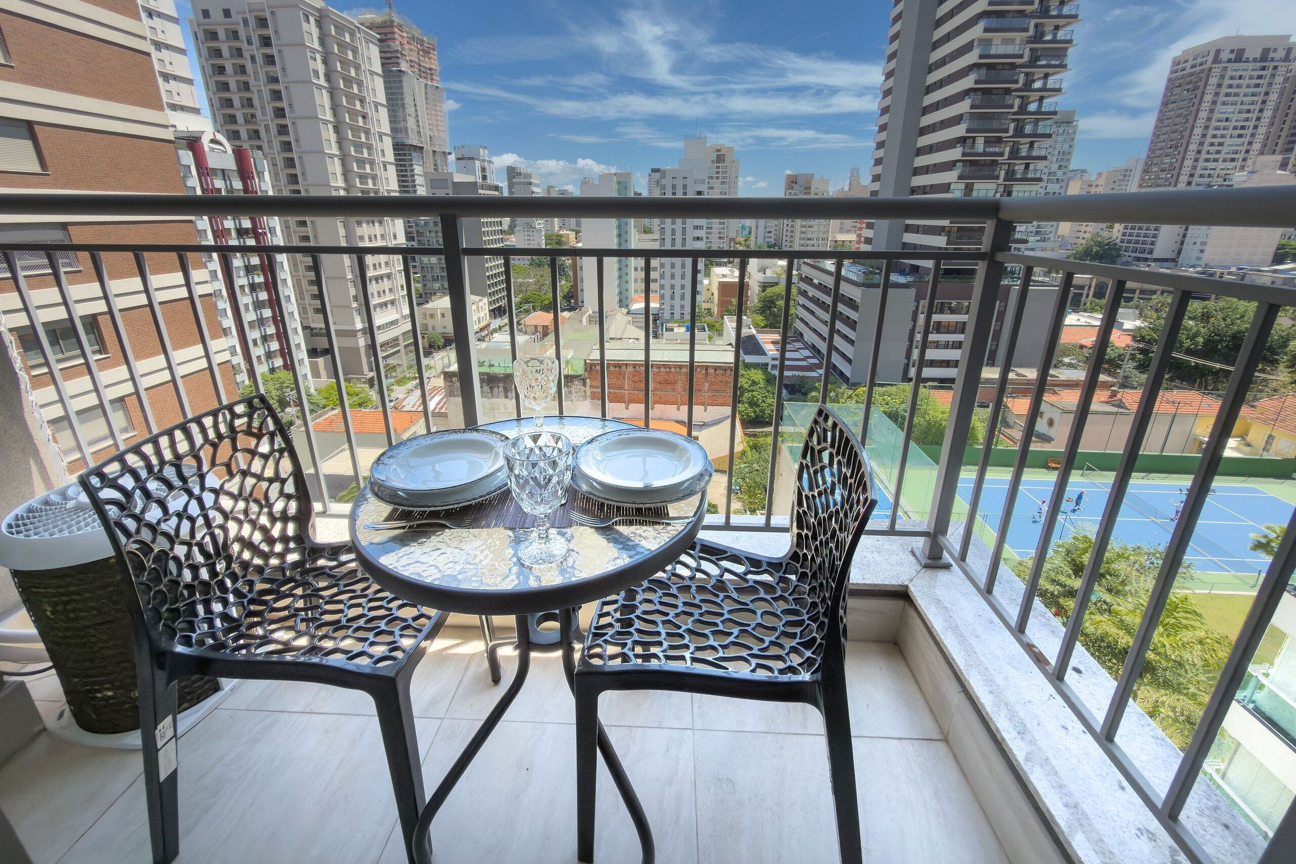 A balcony with a table and chairs, a trash bin, and a view of a cityscape with buildings and a tennis court.