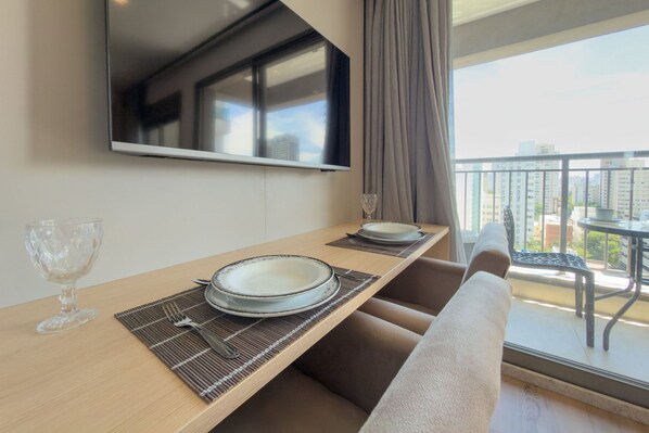 A dining area with a table set for two, a television mounted above, and a balcony with a view of buildings. - Glass Art 96 (São Paulo)