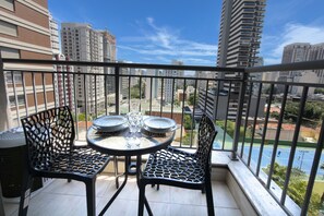 A balcony with a table and chairs, a view of a tennis court, and high-rise buildings in the background. - Glass Art 96 (São Paulo)