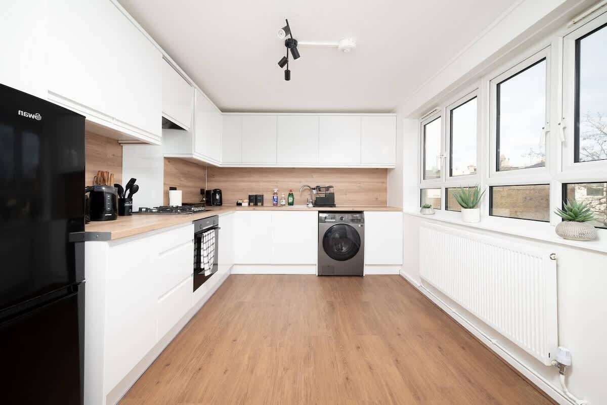 A modern kitchen with white cabinets, a black refrigerator, a built-in washing machine, and a window with plants.