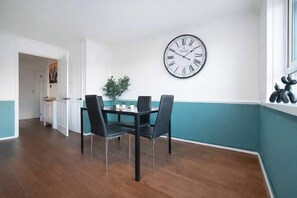 A dining area with a table and chairs, a wall clock, and a window with decorative elements. - Spacious 3 Bed Kensington Flat | BeeMyGuest® (London)