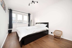 A bedroom with a bed, a window, a wall-mounted light fixture, and a wicker chair. - Spacious 3 Bed Kensington Flat | BeeMyGuest® (London)