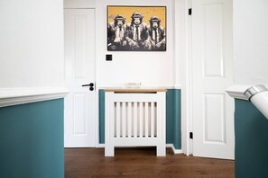 A hallway with a white door, a small white bench with a wooden top, and a painting of three figures on the wall. - Spacious 3 Bed Kensington Flat | BeeMyGuest® (London)
