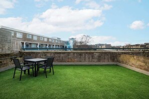 An outdoor patio with a table and chairs, a grassy area, and a view of a building with multiple windows. - Spacious 3 Bed Kensington Flat | BeeMyGuest® (London)